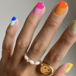 Hand with neon multicolor nails (pink, blue, orange, lime, yellow); wearing a pearl bead ring and a gold smiley-face ring.