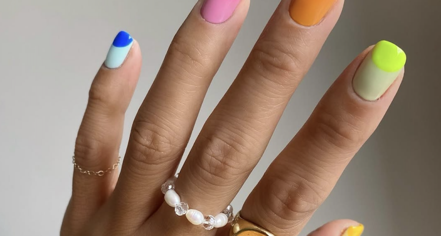 Hand with neon multicolor nails (pink, blue, orange, lime, yellow); wearing a pearl bead ring and a gold smiley-face ring.