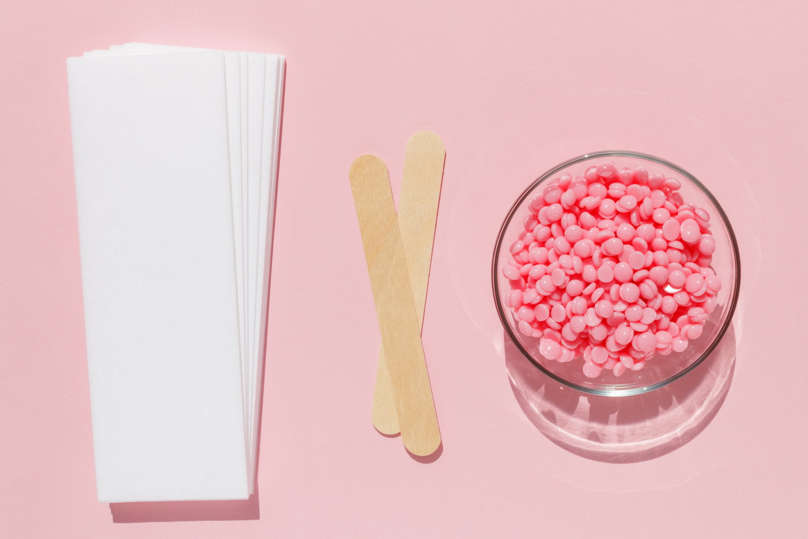 Beautiful pink depilatory wax granules strips for depilation and wooden spatulas on a pink background Epilation depilation unwanted hair removal Top view | Live That Glow Waxing products ready for a Brazilian or bikini wax