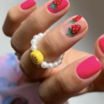 Close-up of a hand with bright pink nails and strawberry nail art, wearing a pearl bracelet with a yellow smiley bead.