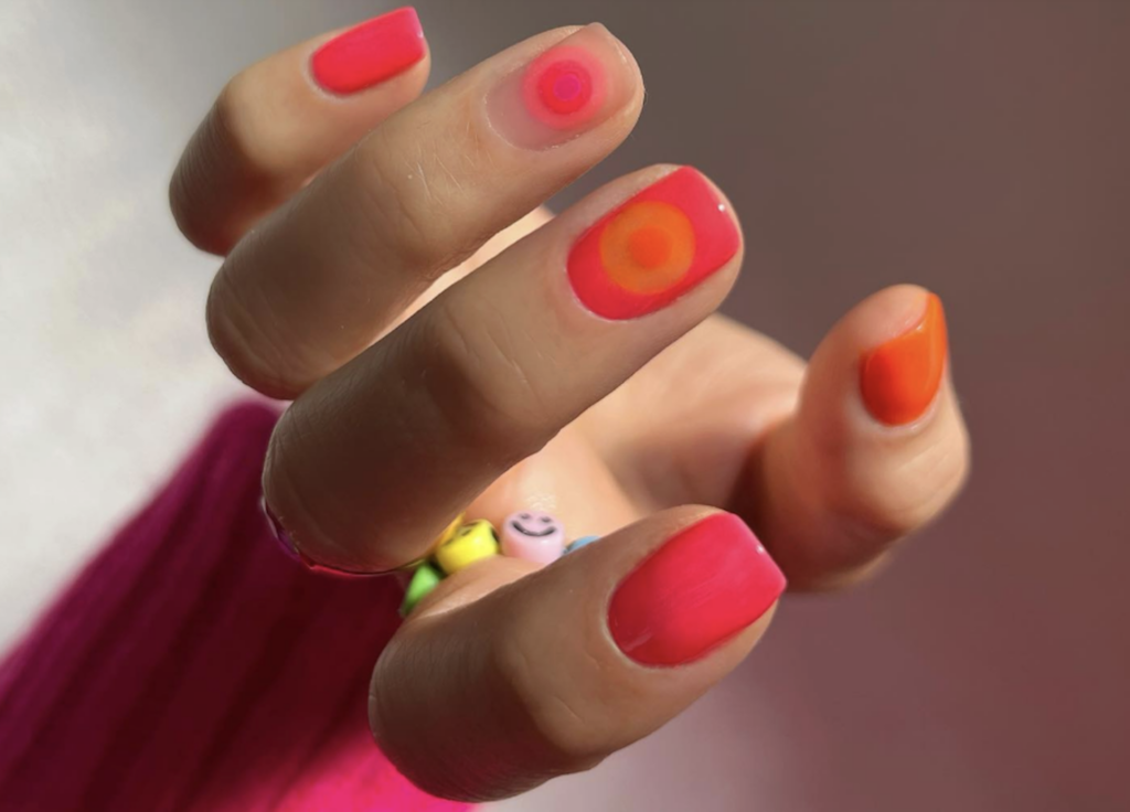 Close-up of a hand with neon pink and orange nails holding a small colorful beaded bracelet with smiley-face beads