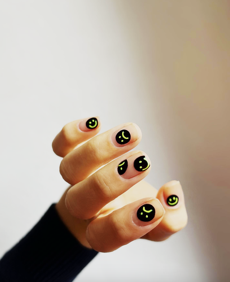 neon-smiley-face-nails-the-hang-edit | Live That Glow Close-up of a hand showing fingernails painted black with neon green smiley faces on each nail.
