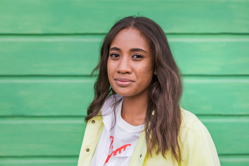 Young Pacific Islander female with long hair and acne looking at camera while standing against green cottage wall in Sonoma County, California