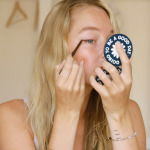 young blonde female, sitting on a bed, in underwear, applying eyeliner on her eye, while looking into a small round mirror which she holds in her hand