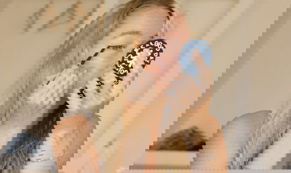 young blonde female, sitting on a bed, in underwear, applying eyeliner on her eye, while looking into a small round mirror which she holds in her hand