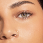 Close-up of a person’s face showing a small gold nose ring and well-groomed brows with warm makeup.