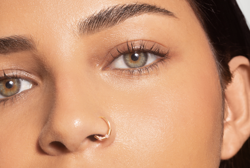 Close-up of a person’s face showing a small gold nose ring and well-groomed brows with warm makeup.