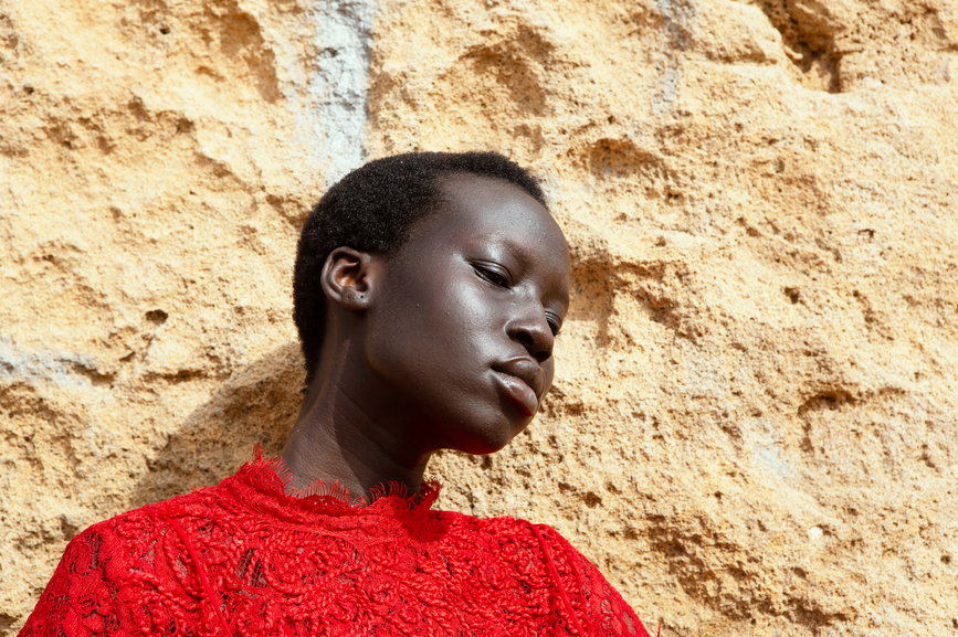 portrait of a young woman of color in a flowing red dress, set against the dramatic backdrop of a Moroccan beach and rock formation. | Live That Glow