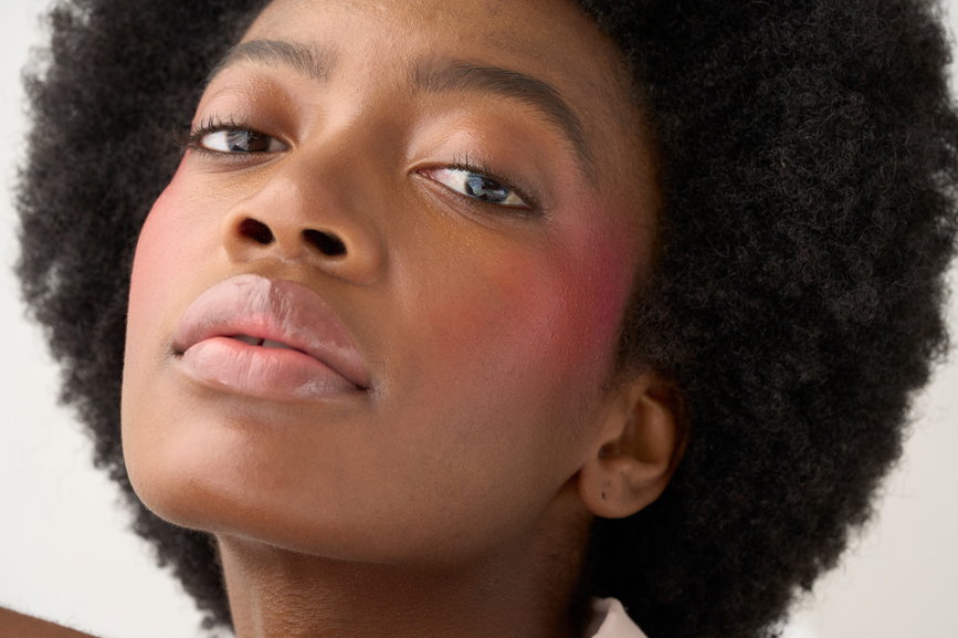 Studio beauty makeup portrait of a woman with a rouge blush on her cheeks and a soft pink shirt and an Afro hairstyle. Neutral background. | Live That Glow