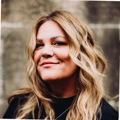 Portrait of a smiling blonde woman with wavy hair, wearing a dark top, outdoors near a stone wall.