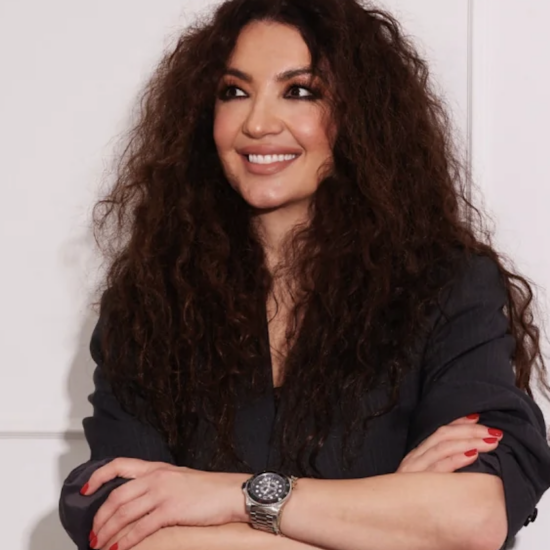 Woman with long curly hair, dark top, smiling with arms crossed, red nails, wearing a silver wristwatch against a pale wall backdrop