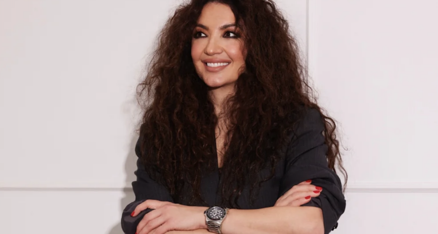 Woman with long curly hair, dark top, smiling with arms crossed, red nails, wearing a silver wristwatch against a pale wall backdrop