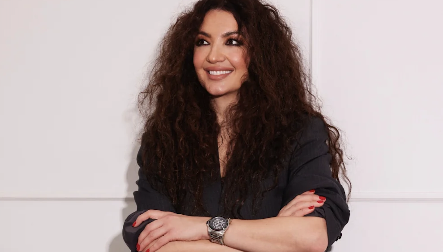 Woman with long curly hair, dark top, smiling with arms crossed, red nails, wearing a silver wristwatch against a pale wall backdrop