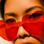 Close-up of a person wearing bright red heart-shaped sunglasses, sunlit face visible behind the lenses