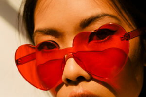 Close-up of a person wearing bright red heart-shaped sunglasses, sunlit face visible behind the lenses