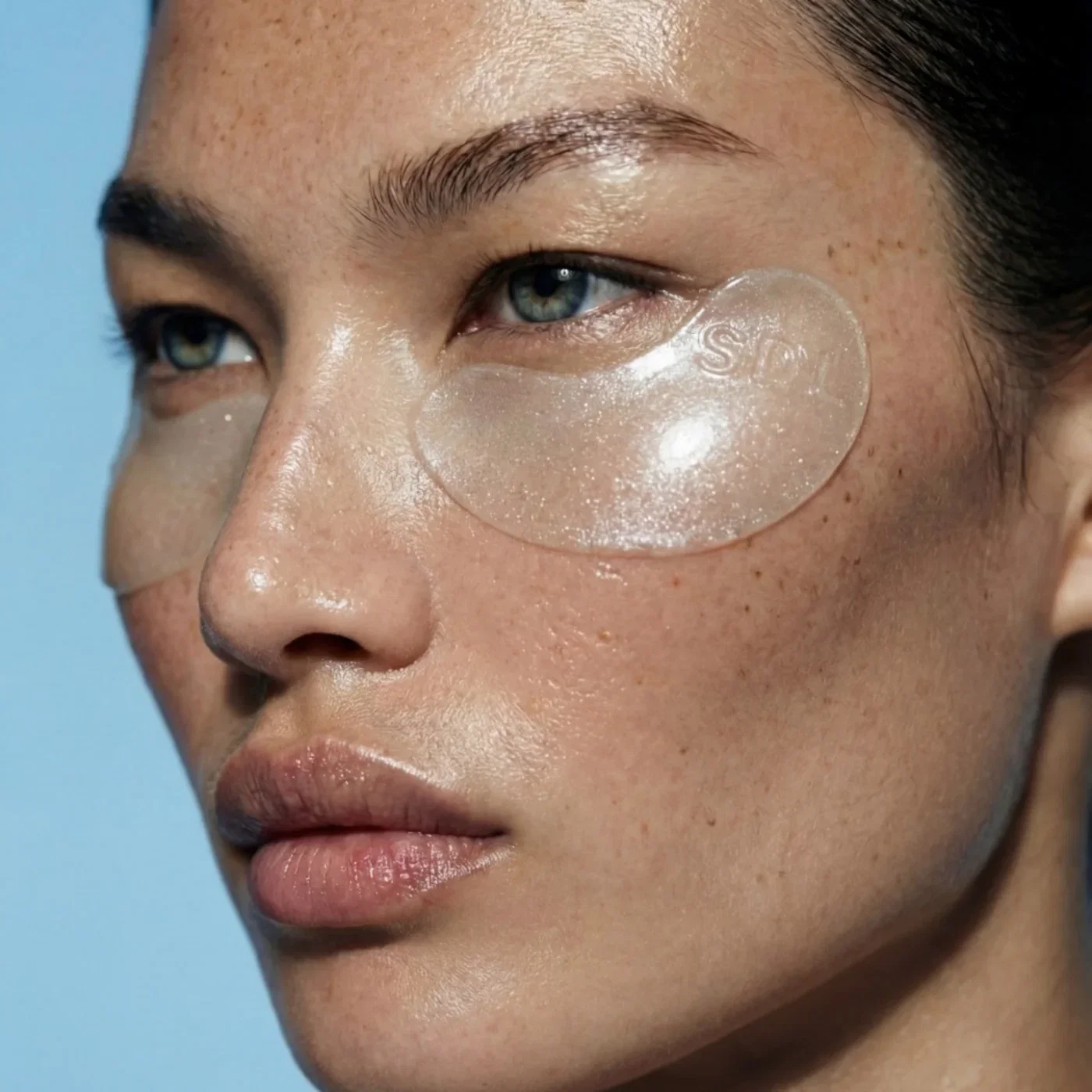Close-up of a woman's face with a translucent under-eye hydrogel patch placed beneath her left eye, blue background.