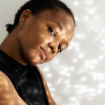 Portrait of a woman in a black sleeveless top, tilting her head with warm, dappled sunlight on her face, looking at the camera.