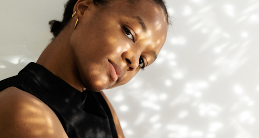 Portrait of a woman in a black sleeveless top, tilting her head with warm, dappled sunlight on her face, looking at the camera.