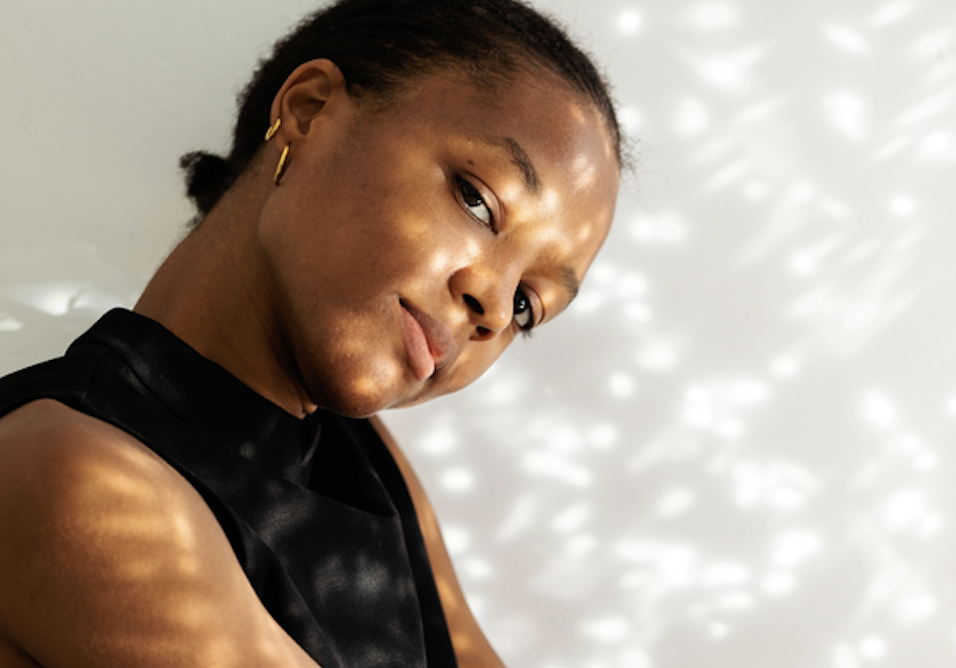 Portrait of a woman in a black sleeveless top, tilting her head with warm, dappled sunlight on her face, looking at the camera.