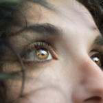 Close-up of a person’s golden-brown eye looking upward, with lashes and soft skin visible and green foliage in the foreground blur.
