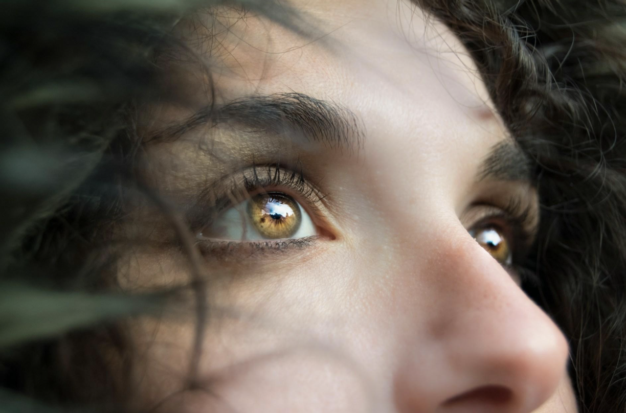 Close-up of a person’s golden-brown eye looking upward, with lashes and soft skin visible and green foliage in the foreground blur.