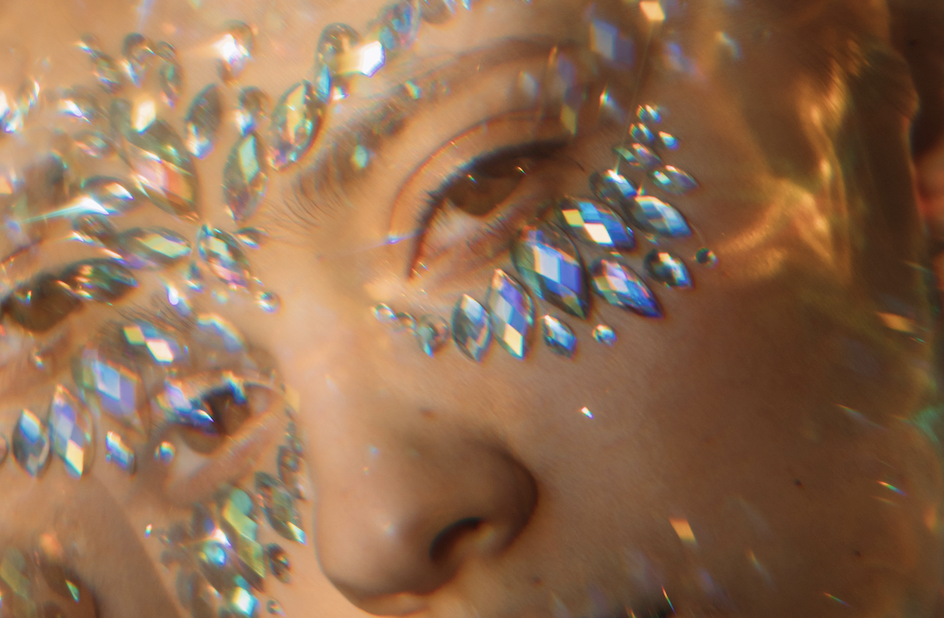 Close-up of a person's eye surrounded by shimmering crystal rhinestones on the cheek and brow, bathed in golden light.