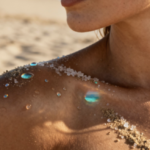 Close-up of a woman's shoulder and neck on a sunny beach, with sand grains and iridescent droplets clinging to her skin.