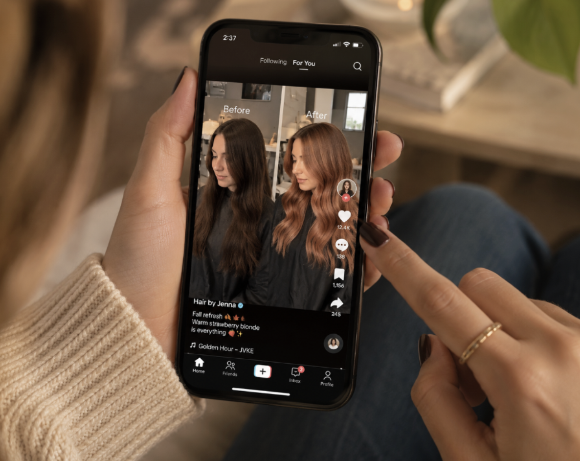 A person holding a smartphone showing a TikTok video with a before/after hair transformation, blonde waves on the right and brown hair on the left.