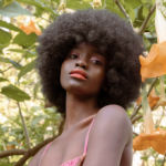 Portrait of a woman with a large Afro among orange hanging flowers in a sunlit garden.