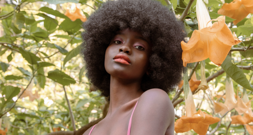 Portrait of a woman with a large Afro among orange hanging flowers in a sunlit garden.