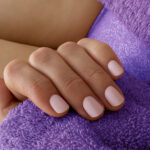 Close-up of a hand with pale pink manicured nails resting on a soft purple plush blanket.