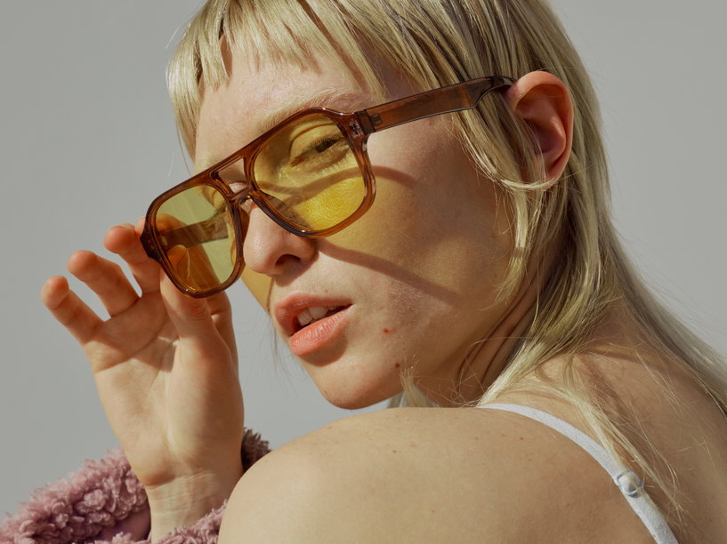 Woman wearing oversized amber sunglasses, looking over her shoulder, with blonde hair and a soft pink coat draped nearby.