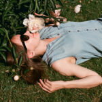 Woman lying on grass among blooming pink peonies, wearing a light denim sleeveless jumpsuit and sunglasses, smelling a flower.