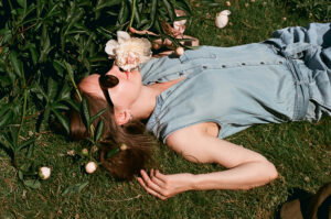Woman lying on grass among blooming pink peonies, wearing a light denim sleeveless jumpsuit and sunglasses, smelling a flower.