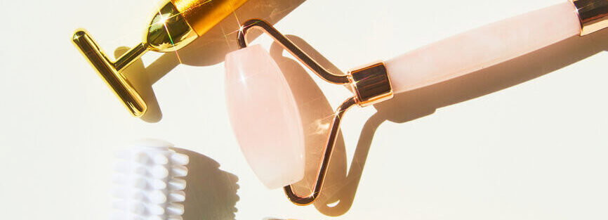 Set of vintage-style grooming tools on a light background: gold-toned razor, pink-handled razor with curved metal frame, and a white bristle brush.