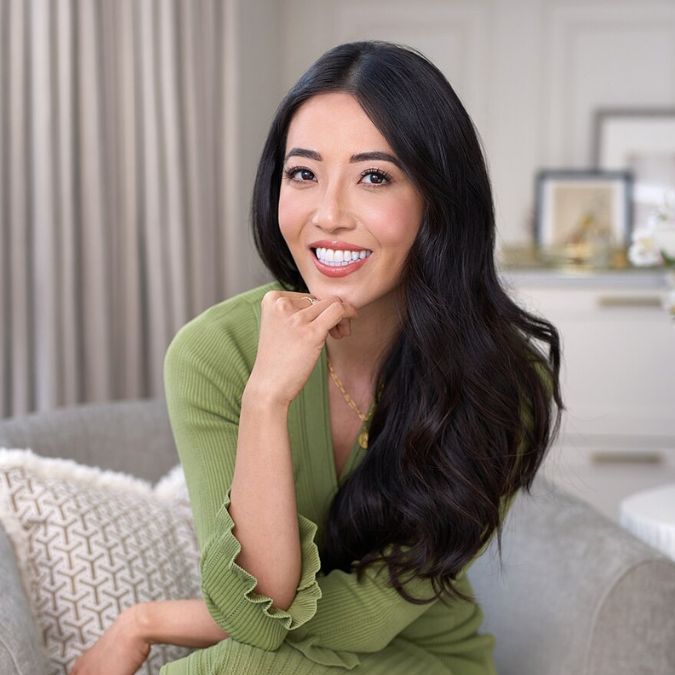 Susie-Ma | Live That Glow Smiling woman with long dark hair in a green sweater sits on a sofa in a cozy living room/mentions her relaxed pose with her hand near her chin.