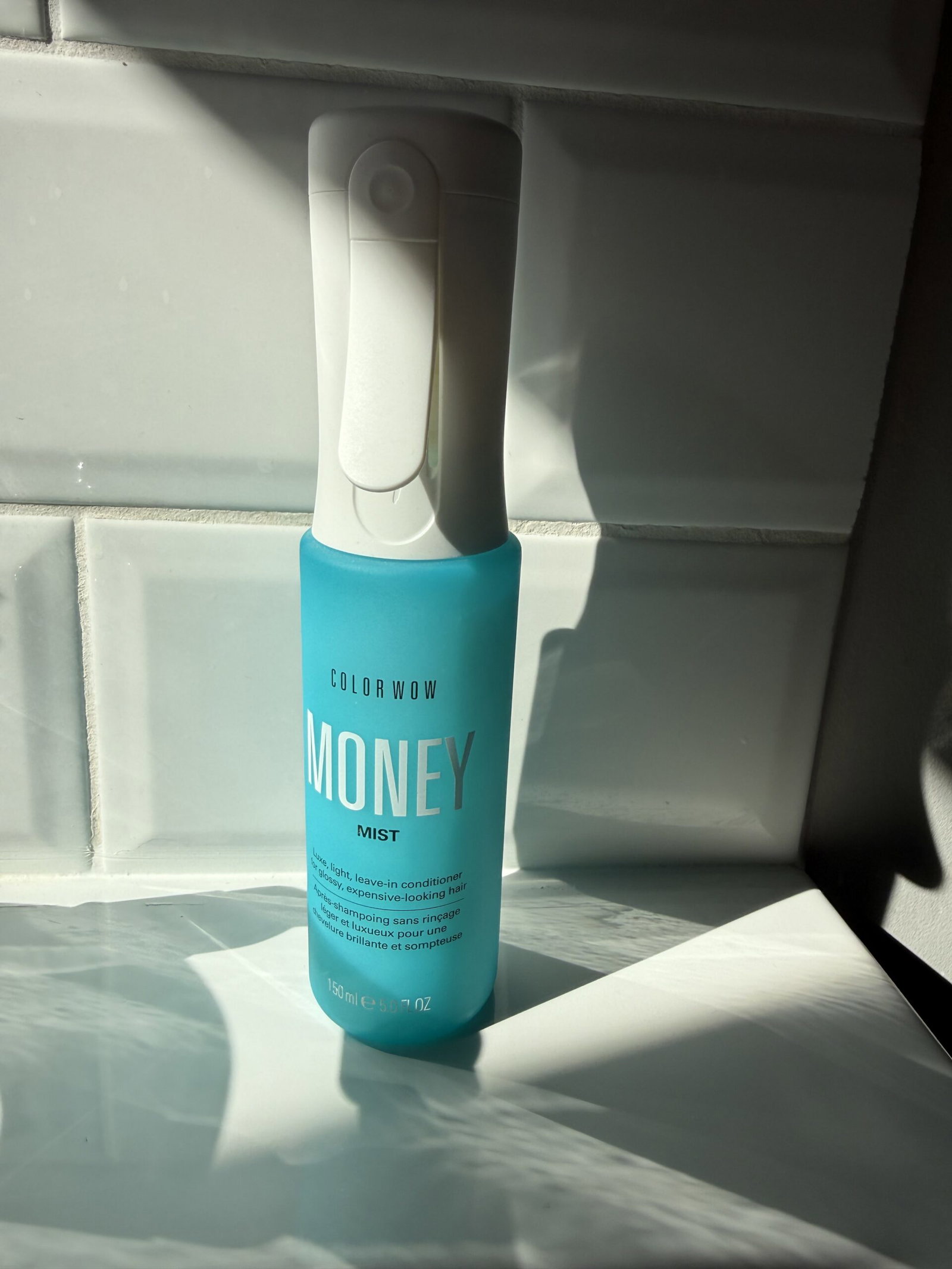 Turquoise bottle of Color Wow Money Mist sitting on a white windowsill in bright sunlight.