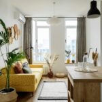 Bright open-plan living room with a yellow sofa, green plants, woven wall baskets, and large windows letting in light.