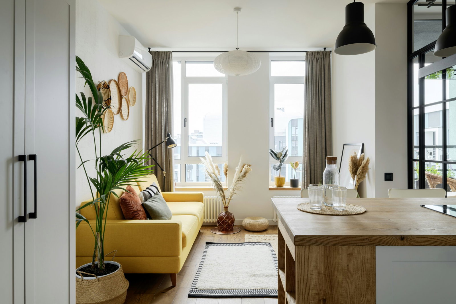 Bright open-plan living room with a yellow sofa, green plants, woven wall baskets, and large windows letting in light.