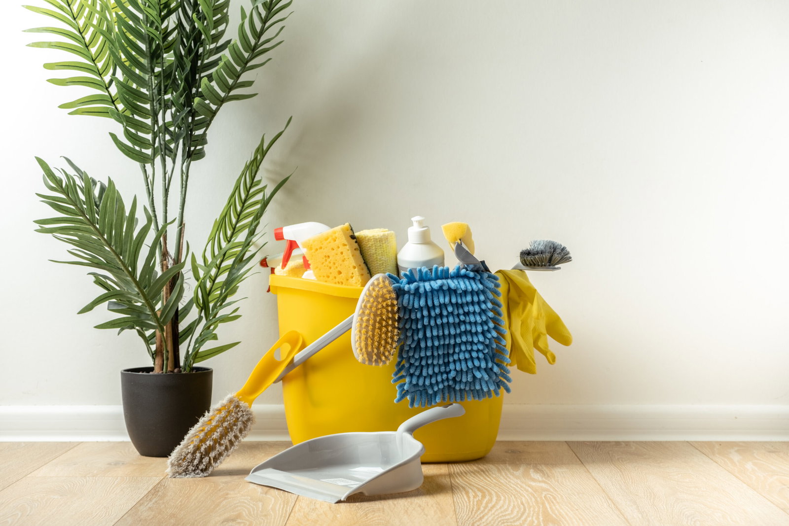 Yellow cleaning bucket overflowing with sponges, brushes, gloves, and a spray bottle beside a houseplant on a wooden floor.