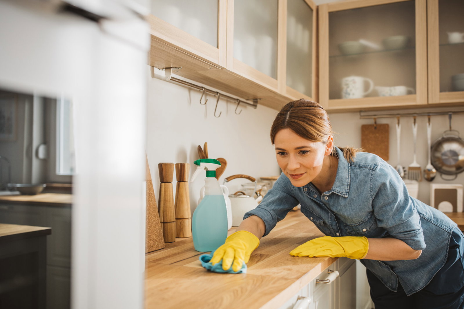Woman in a denim shirt wearing yellow gloves wipes the kitchen counter with a spray bottle nearby.