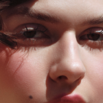 Close-up portrait of a young woman outdoors, with sunlit skin and pink cheeks, focusing on her eyes and nose.
