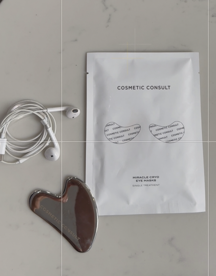 | Live That Glow White cosmetic consultation package on a light marble surface, beside a brown heart-shaped eye mask and a tangled pair of white earphones.
