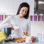 Woman blending a smoothie in a kitchen, pouring milk into a blender while holding a cup with fruit pieces on the counter