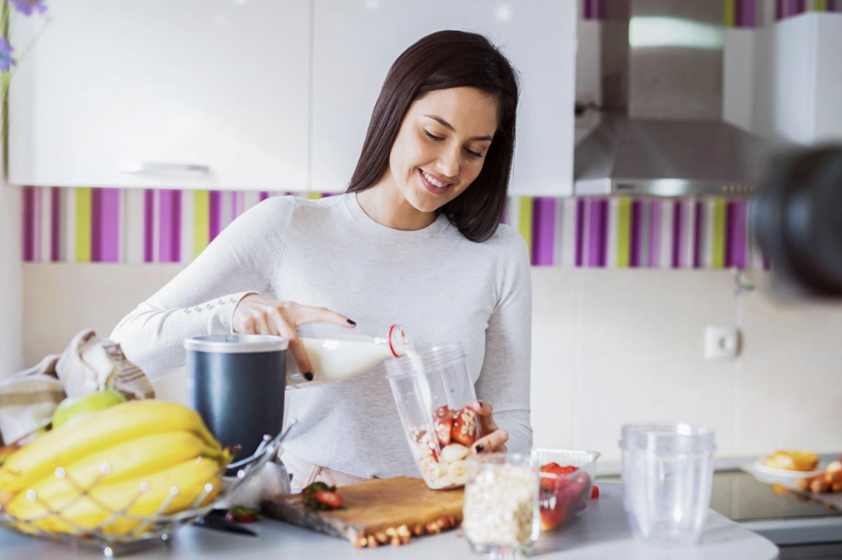 Woman blending a smoothie in a kitchen, pouring milk into a blender while holding a cup with fruit pieces on the counter