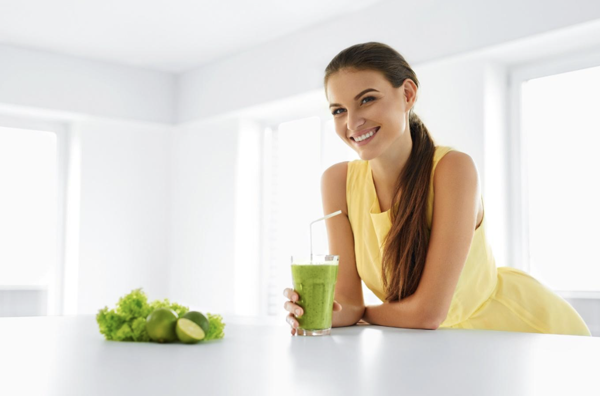 Smiling woman in a yellow dress leans on a white kitchen counter, holding a green smoothie with a straw; fresh greens and limes on the counter.