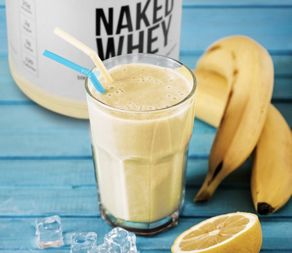 Banana smoothie in a glass with two straws on a bright blue wooden surface, with bananas, lemon half, ice cubes, and a protein powder container in the background.