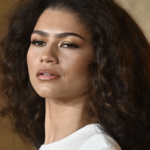 Portrait of a woman with curly dark hair and gold eyeshadow looking slightly upward, against a warm background.