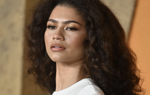Portrait of a woman with curly dark hair and gold eyeshadow looking slightly upward, against a warm background.