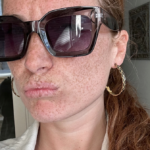 Close-up portrait of a freckled person wearing oversized black sunglasses and a gold hoop earring, with their red hair pulled back in a ponytail.